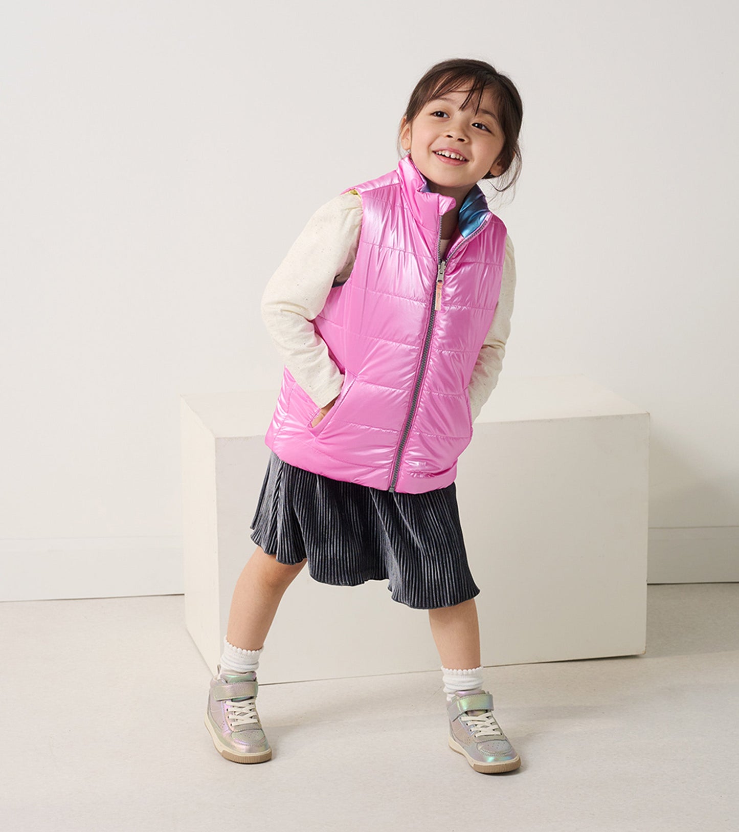 Reversible quilted vest in shiny pink, featuring a high collar and side pockets, worn over a light sweater and skirt.