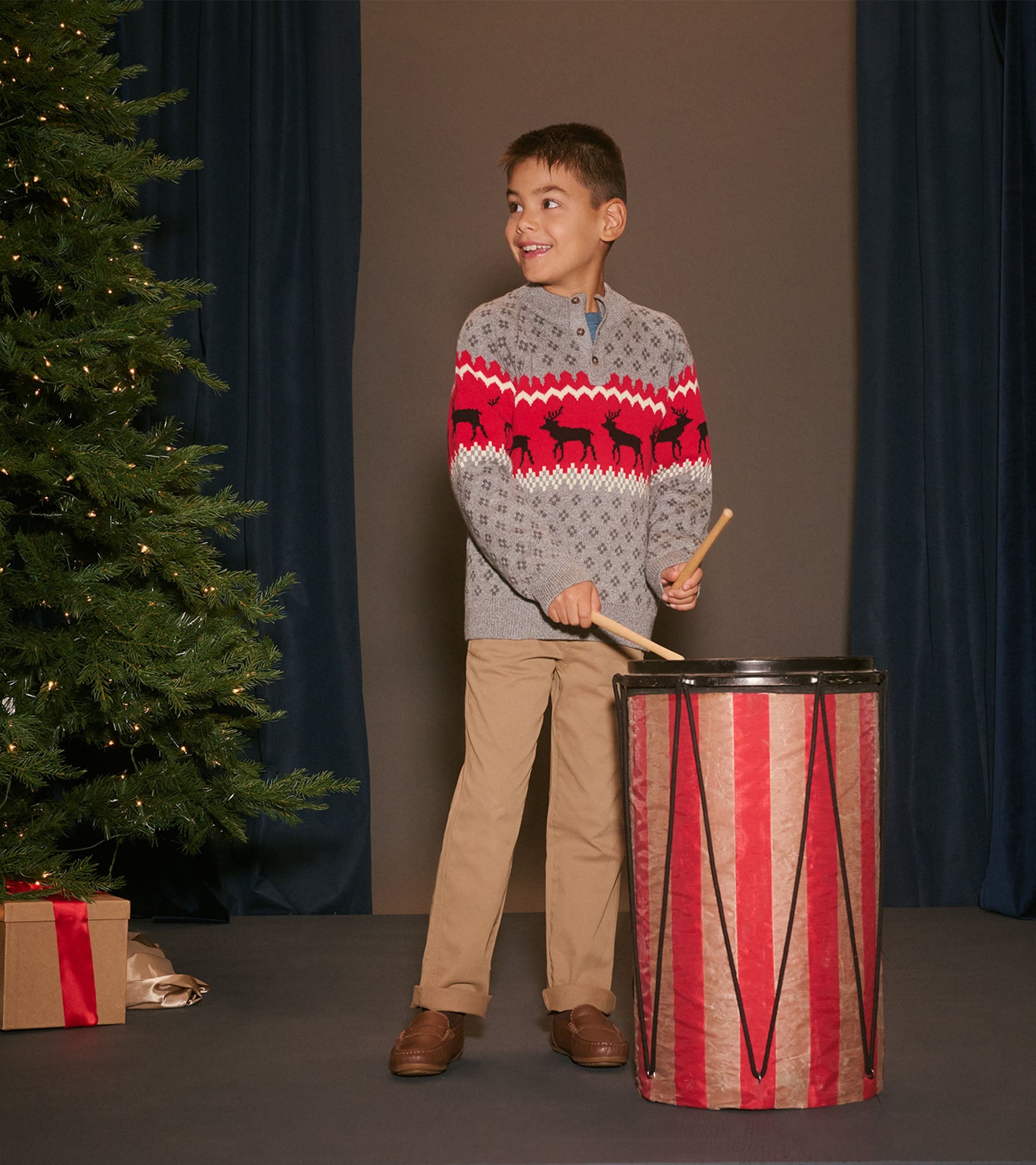 Gray mockneck sweater with red and black elk pattern, worn by a boy playing a drum, festive holiday setting.