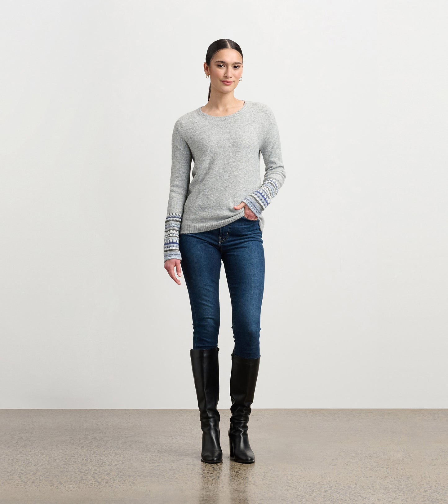 Grey melange sweater with patterned cuffs, crew neck, and relaxed fit, paired with dark jeans and knee-high boots.