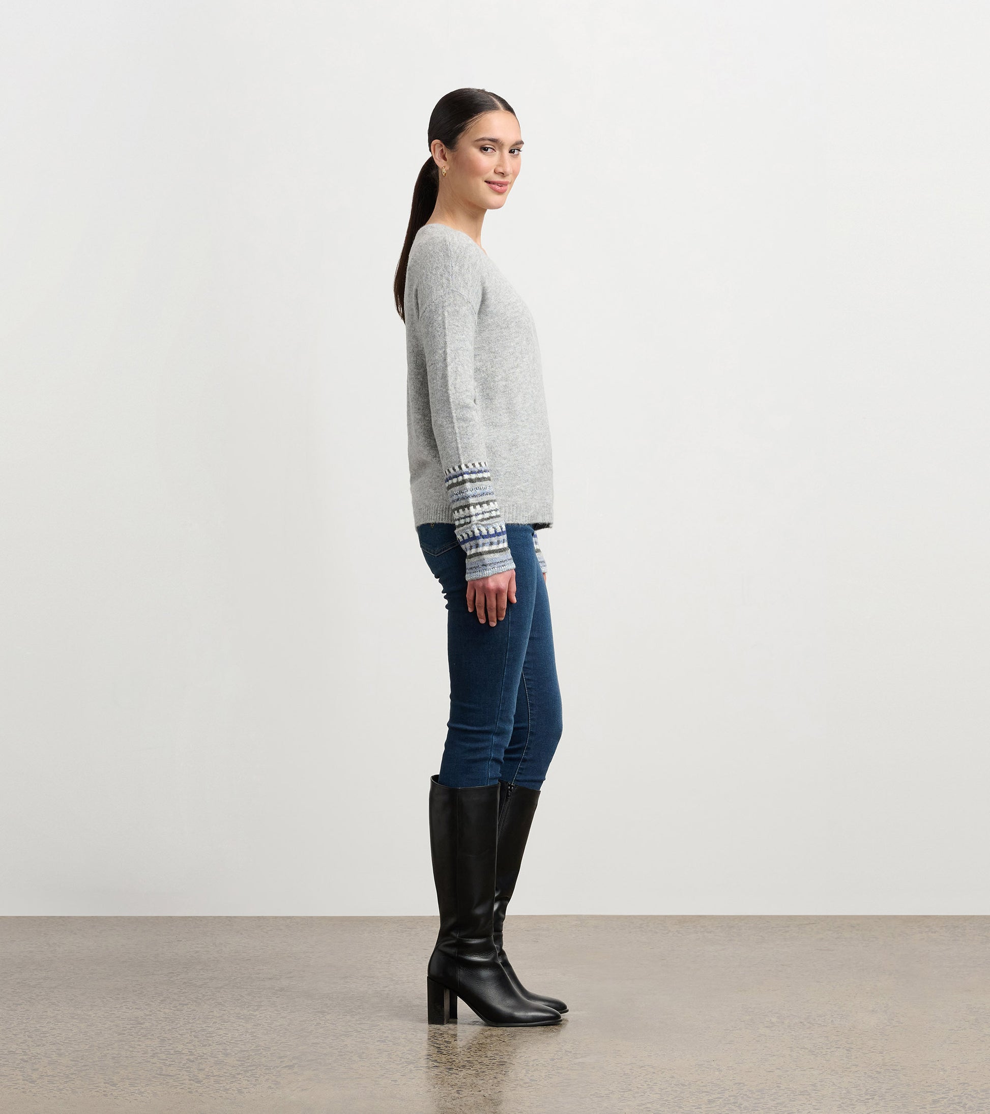 Grey melange sweater with patterned cuffs, featuring a relaxed fit and long sleeves, styled with jeans and knee-high boots.