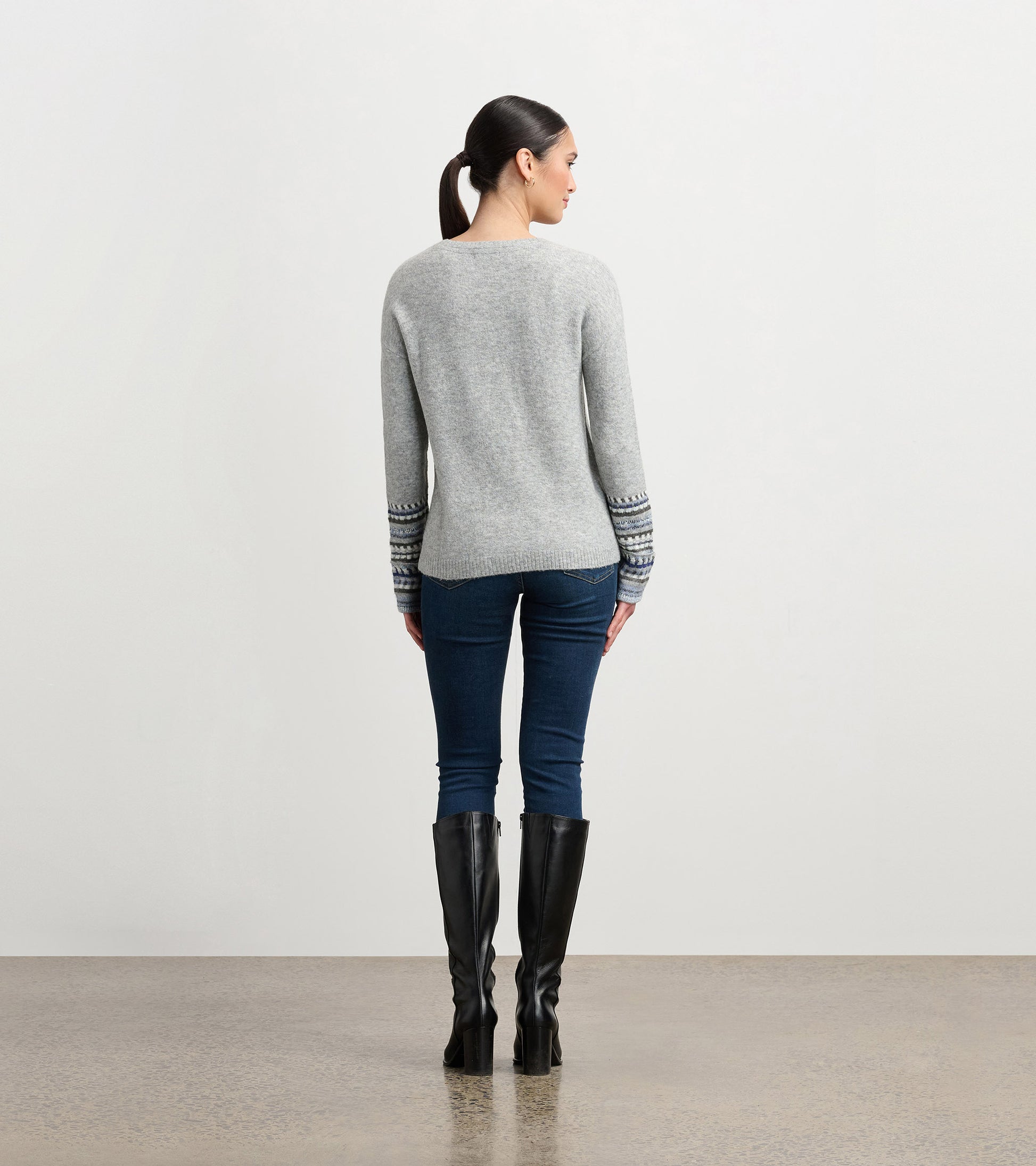 Grey melange sweater with patterned cuffs, fitted shape, and long sleeves, worn with dark jeans and knee-high boots.