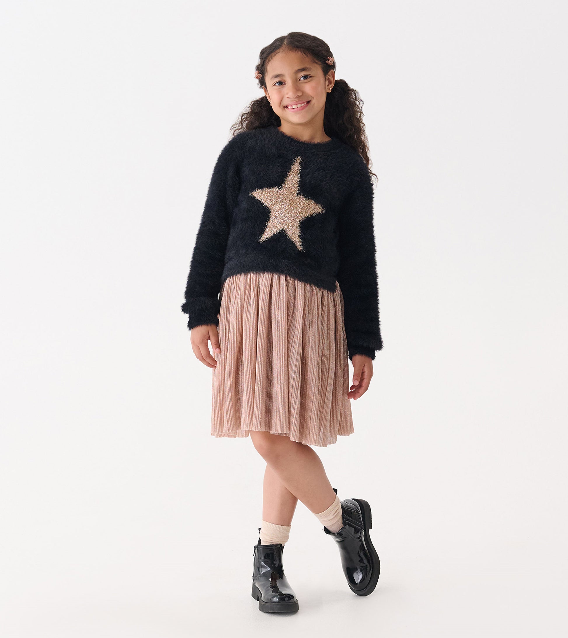 Black fuzzy sweater with a gold star, paired with a pleated pink skirt and black ankle boots.