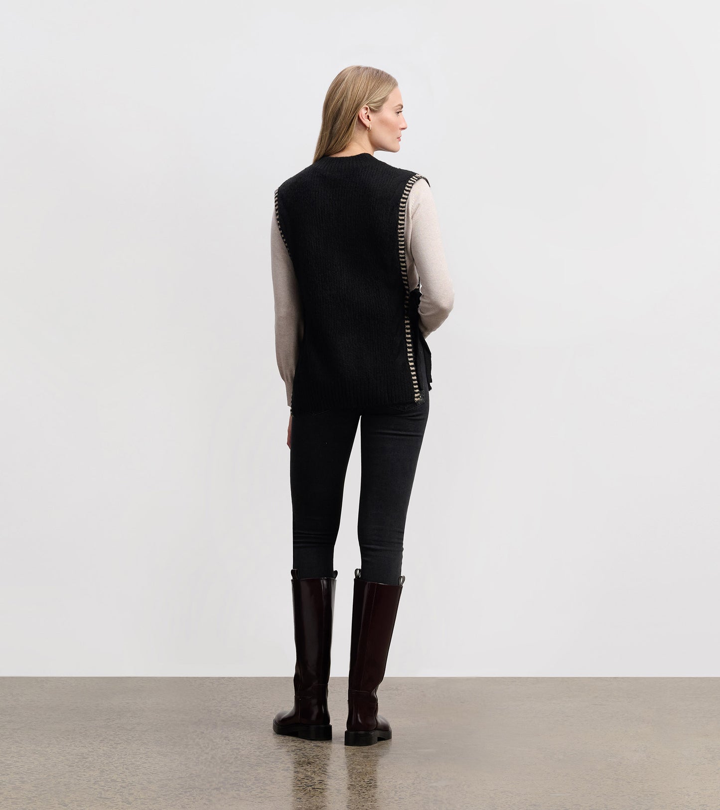 Black side-tie sweater vest with a textured finish, styled over a light top, paired with dark jeans and knee-high boots.