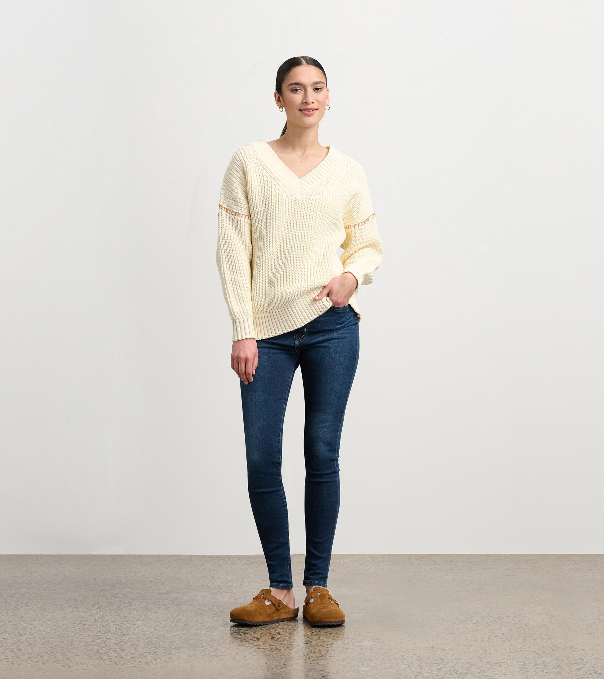 Cream V-neck slouchy sweater with ribbed texture, paired with dark jeans and brown clogs, styled casually.