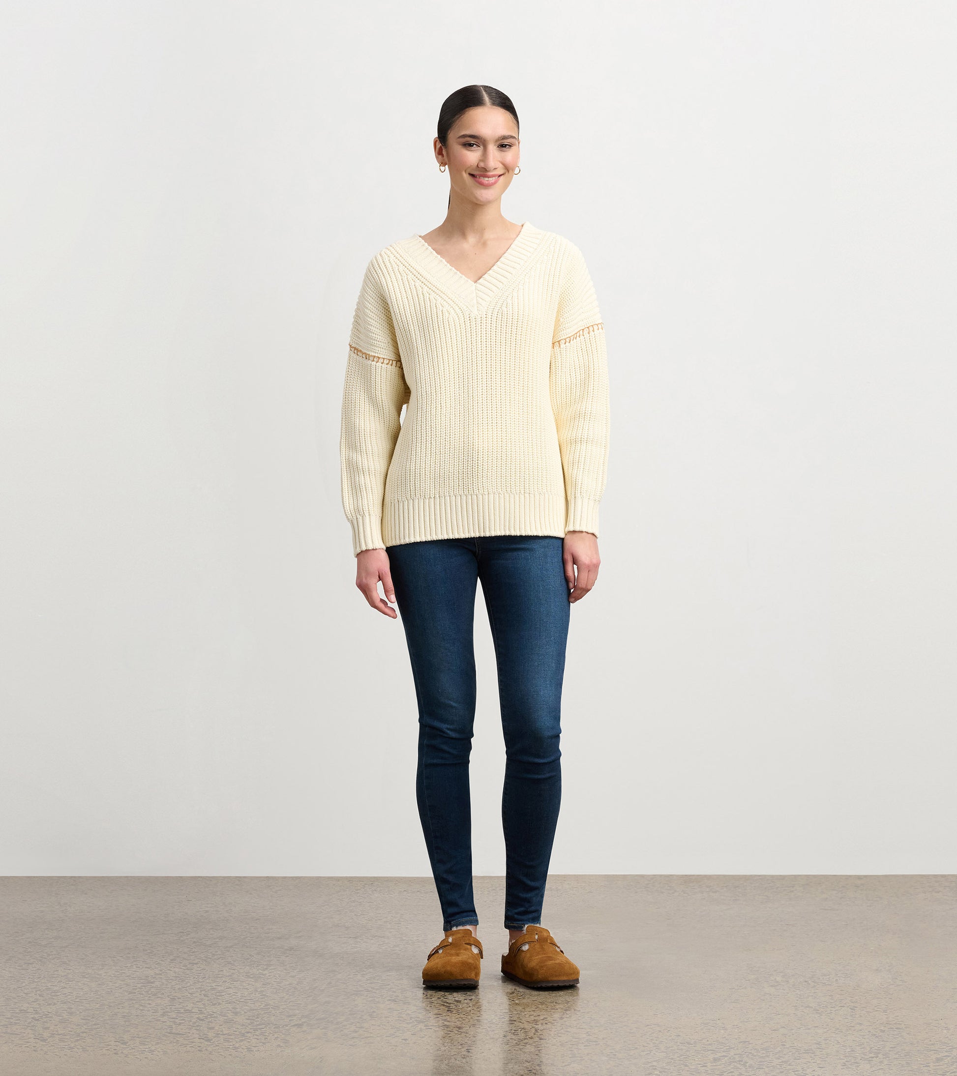 Cream V-neck slouchy sweater with ribbed texture and relaxed fit, paired with dark jeans and brown shoes.