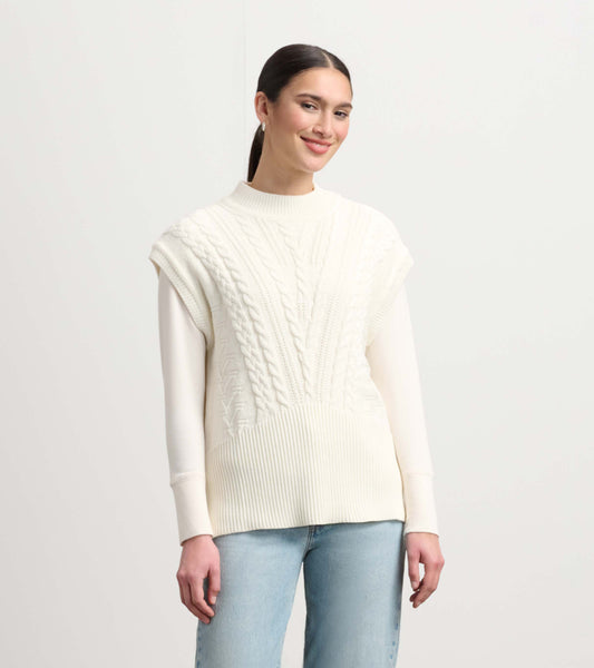 Cream cable knit sweater vest with a high neckline and short sleeves, styled over a long-sleeve top.