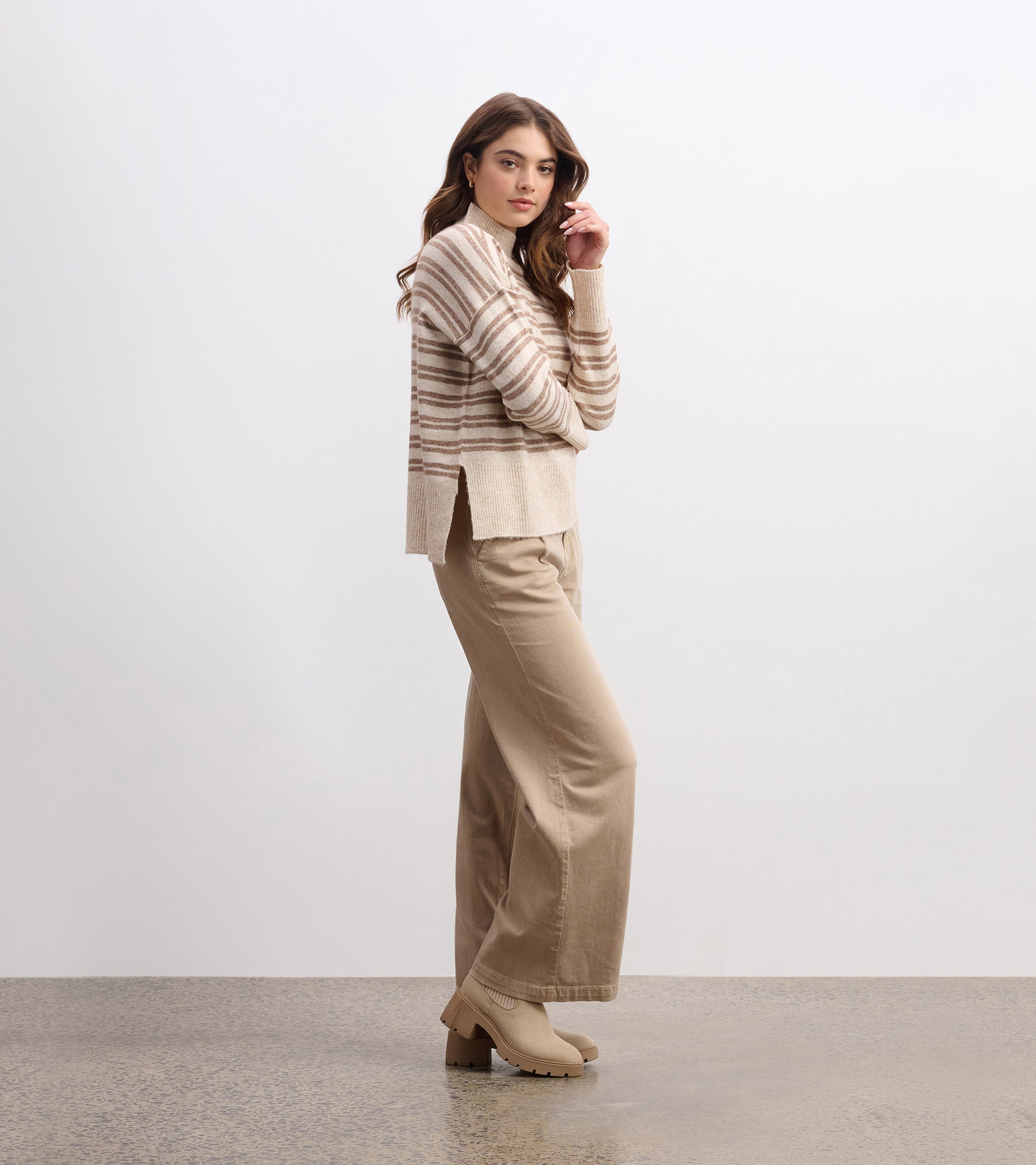Mock neck sweater in camel with double stripes, featuring a relaxed fit and side slits, paired with beige wide-leg pants.