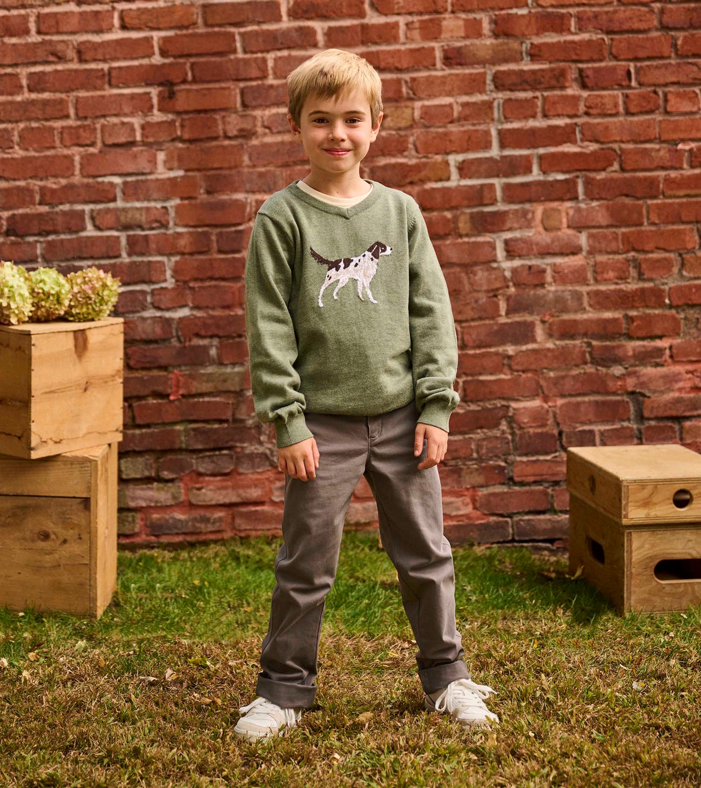 Green V-neck sweater featuring an embroidered hunting dog design, worn by a boy standing against a brick wall.