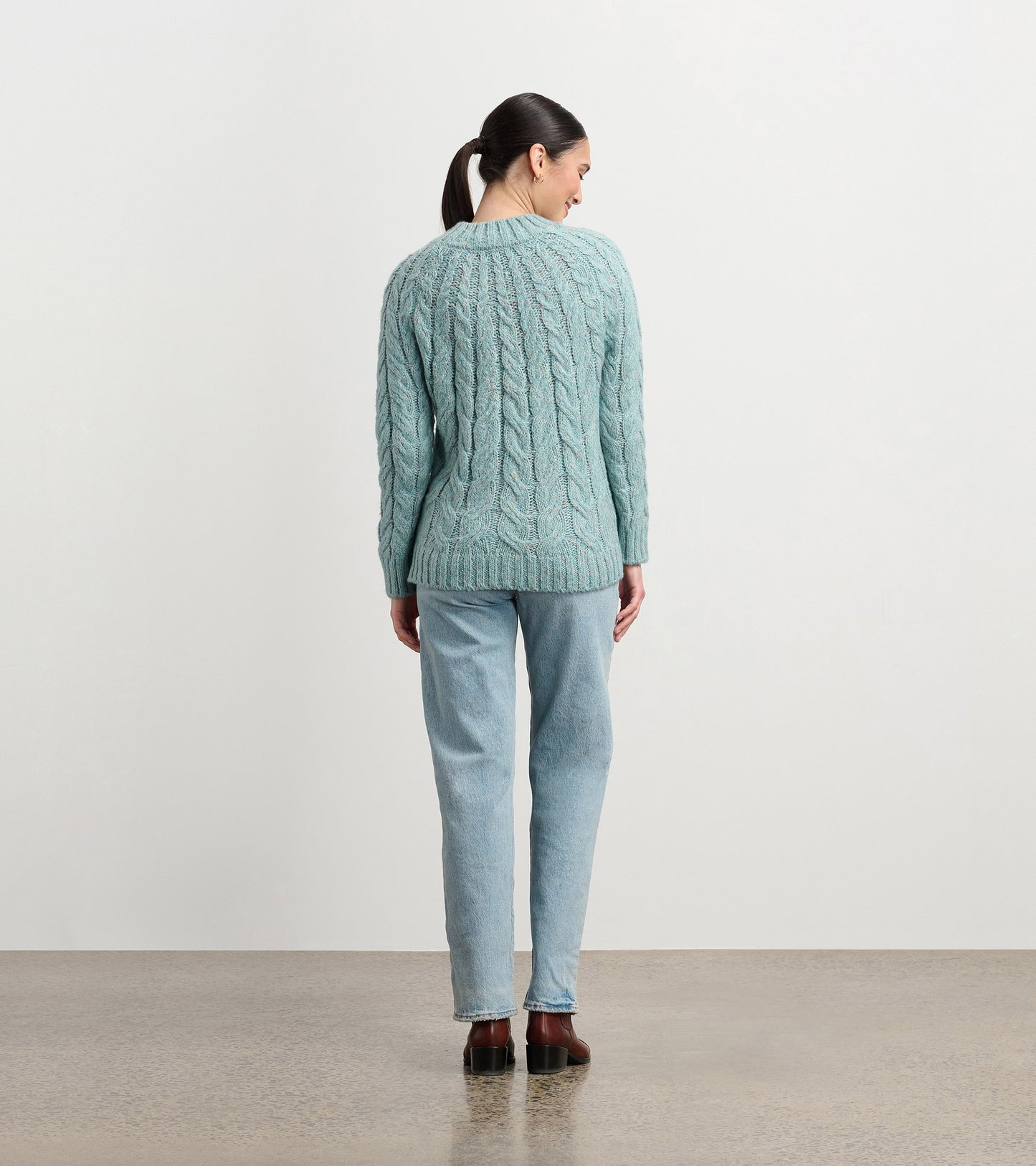 Back view of a light turquoise cable knit sweater with long sleeves and ribbed hem, worn with jeans.