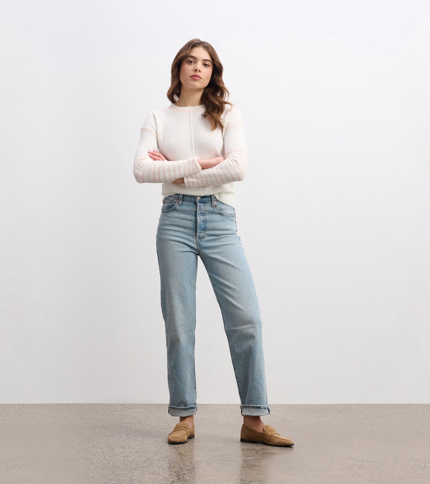 Cream sweater with subtle striped sleeves, paired with light blue jeans and loafers, standing with arms crossed.