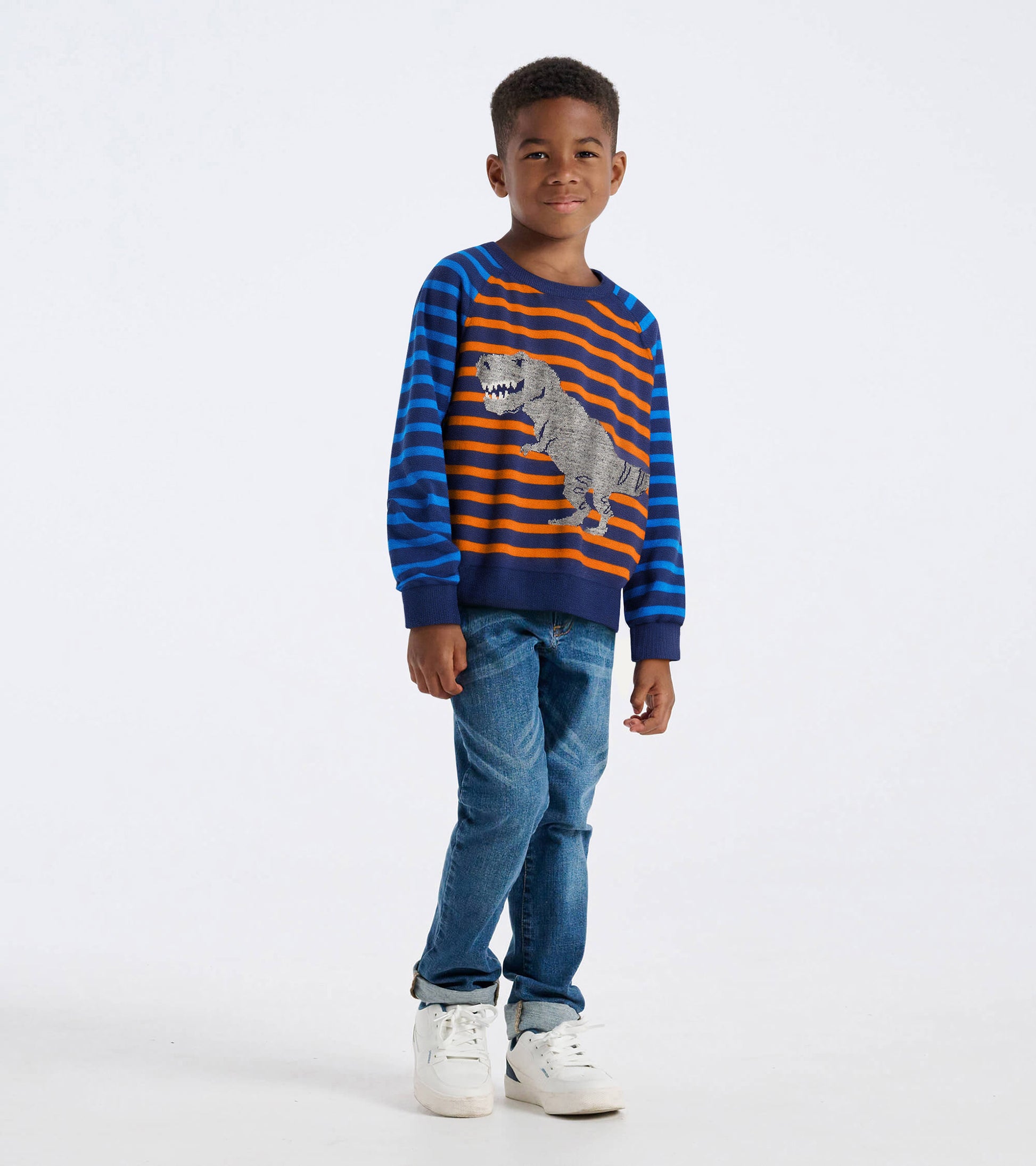 Blue and orange striped raglan sweater featuring a dinosaur graphic, worn by a boy with jeans and sneakers.