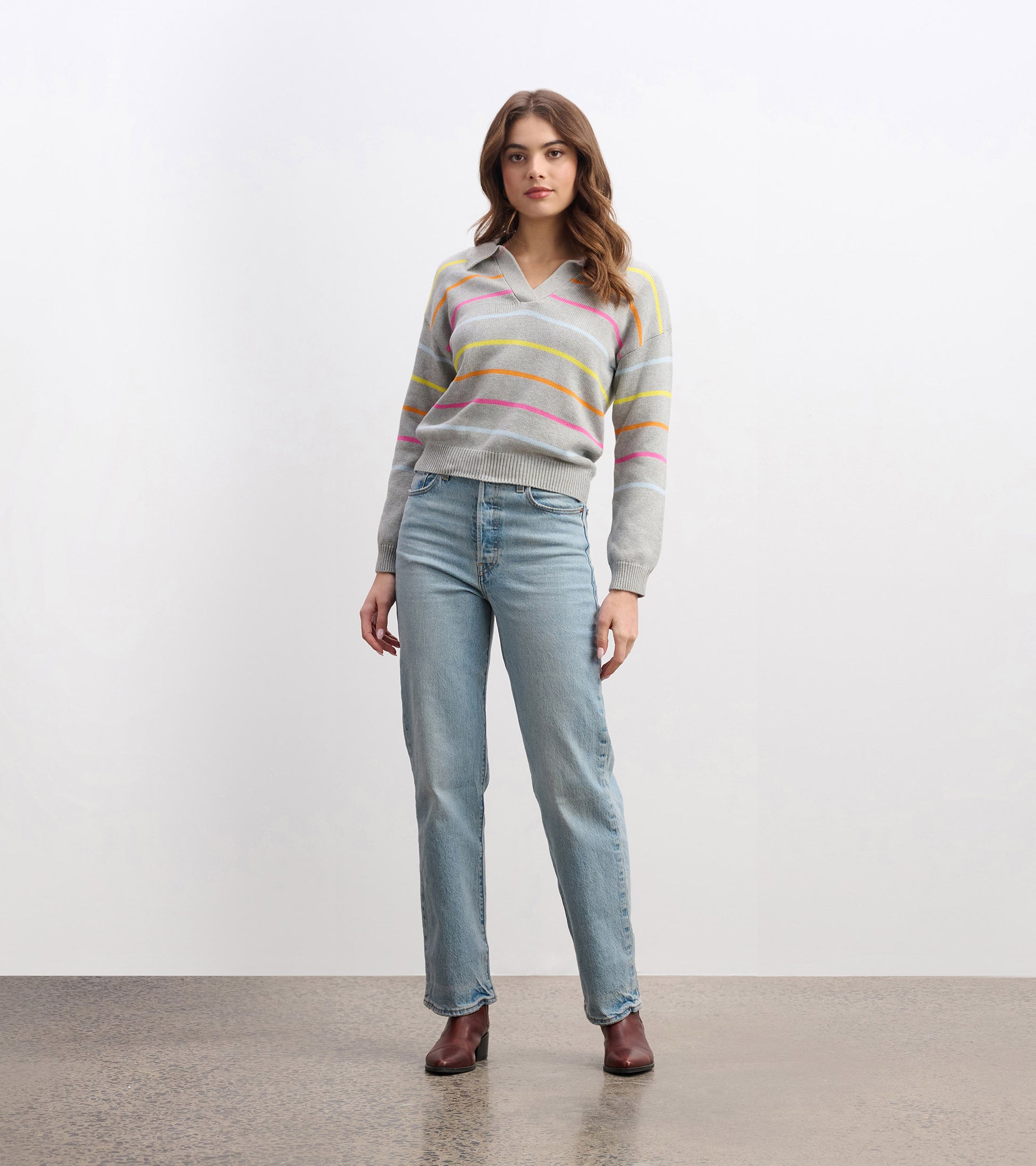 Gray v-neck sweater with colorful stripes, paired with light blue jeans and brown boots, standing against a neutral backgrou…