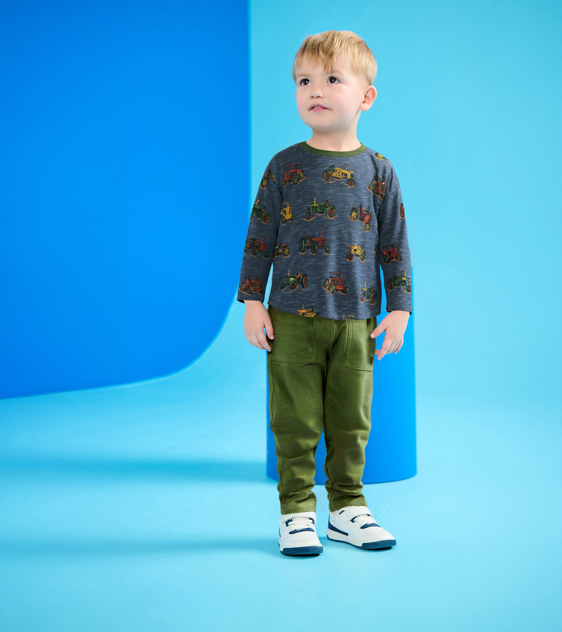 Gray long-sleeve top with tractor print and green pants, worn by a boy standing against a blue background.