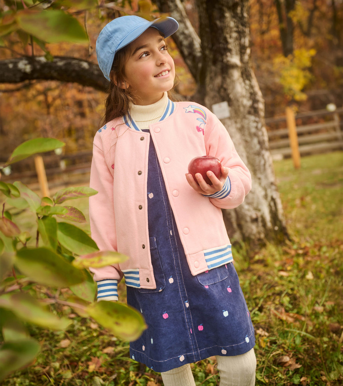 Girl wearing navy apple embroidered pinafore dress under a pink jacket, holding a red apple outdoors