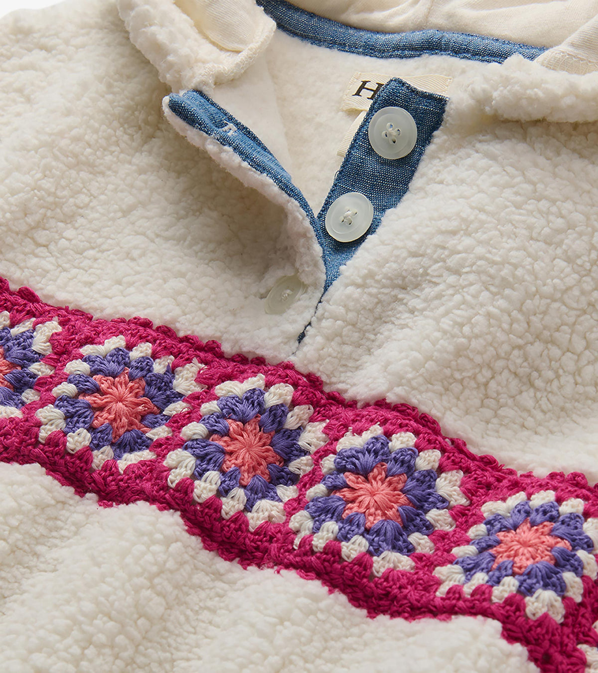 Close-up of cream pullover with blue placket, white buttons, and pink, purple, and coral crochet band