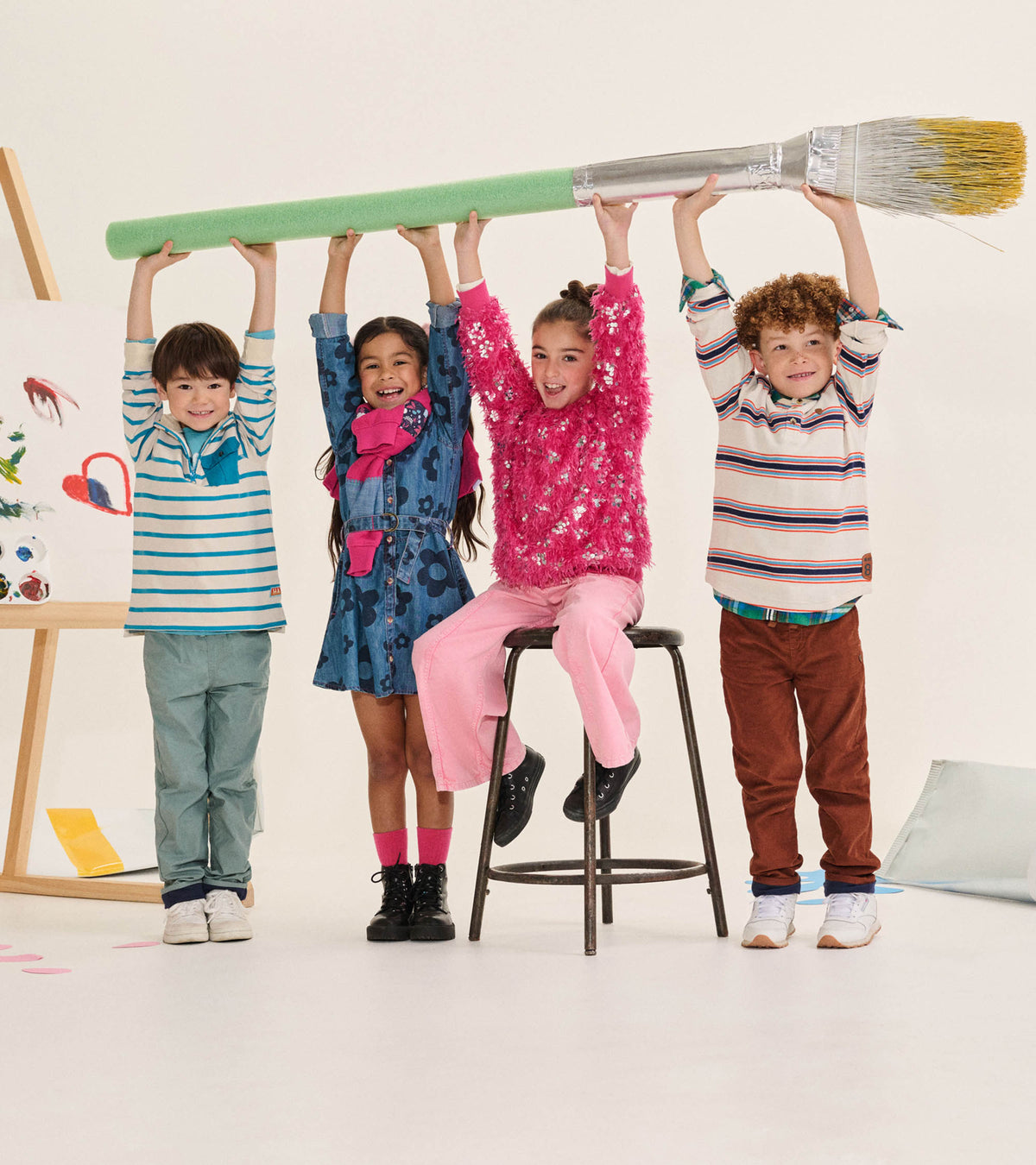 Girl wearing flamingo pink wide leg jeans, seated on a stool with kids holding an oversized paintbrush