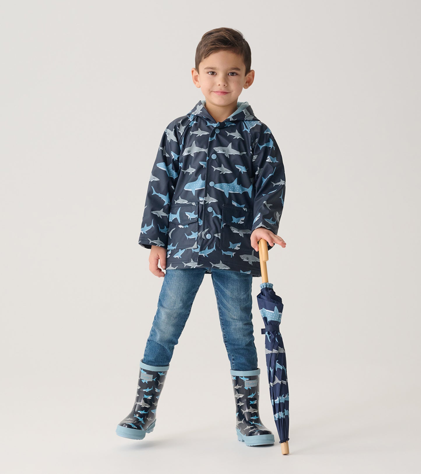 Child in dark blue shark-print raincoat and matching rain boots, holding a folded shark-print umbrella