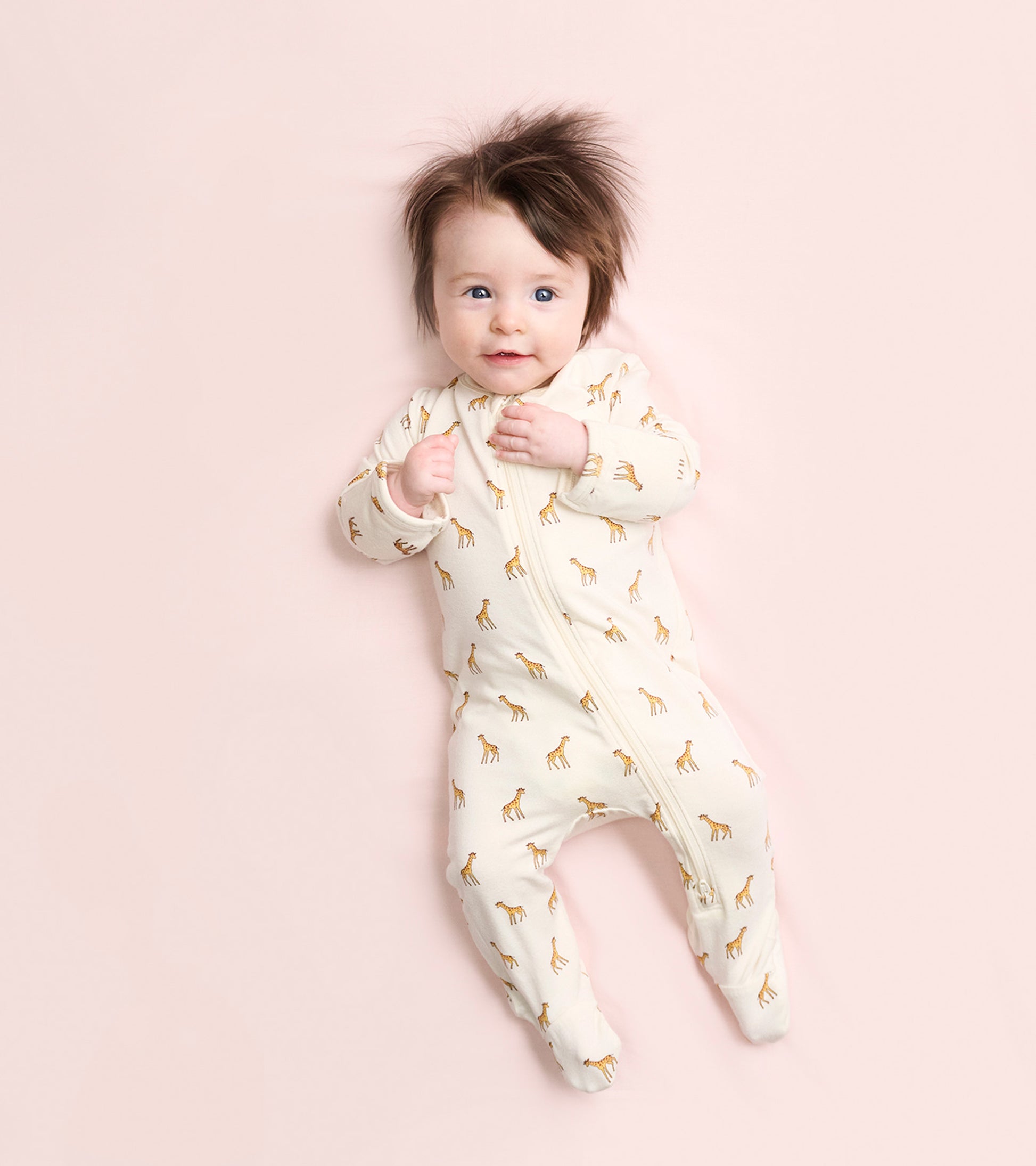 Cream footed sleeper with giraffe print, featuring a front zipper and long sleeves, worn by a smiling baby.