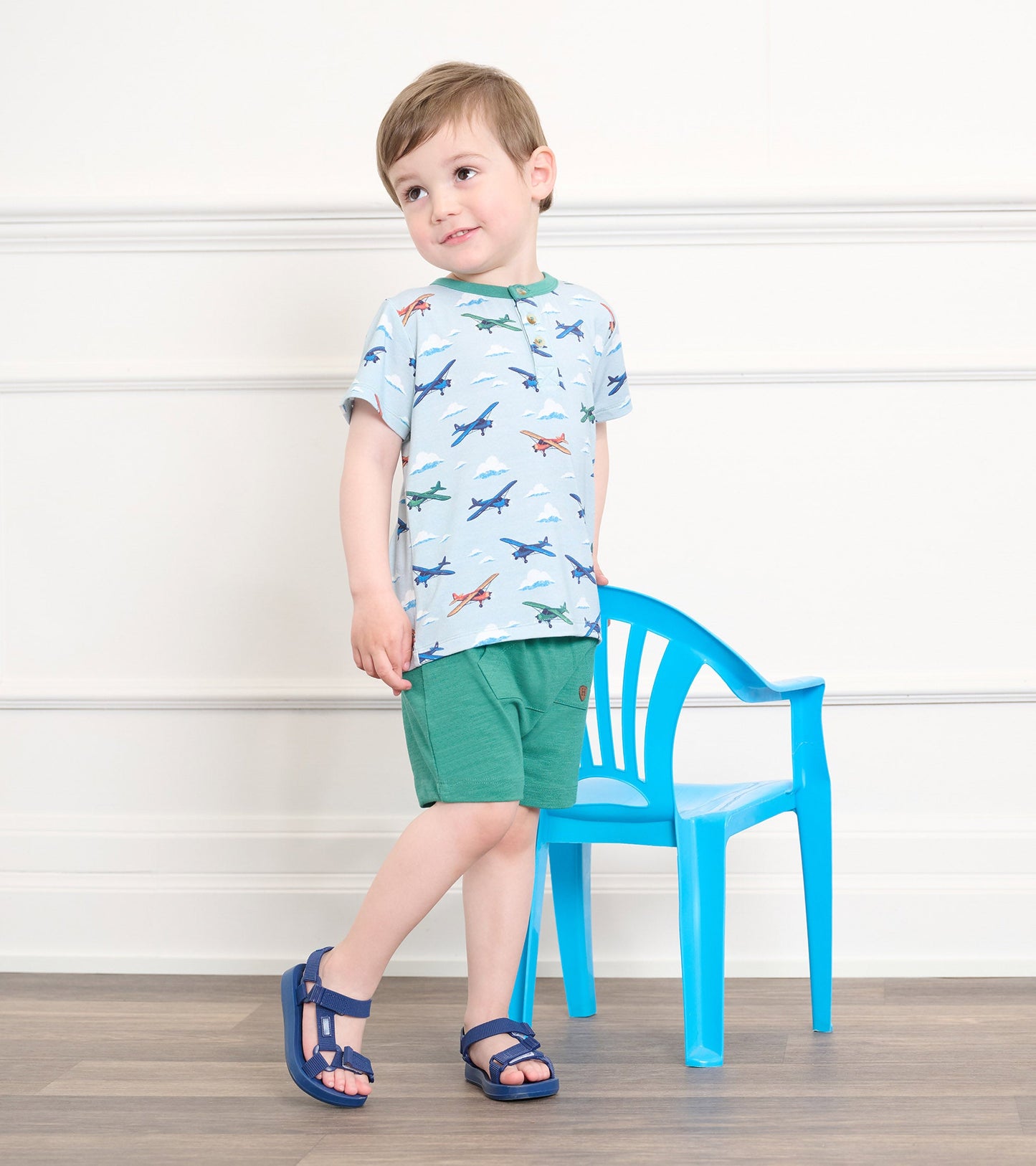Light blue bamboo henley with airplane print, paired with green shorts, worn by a boy standing next to a blue plastic chair.