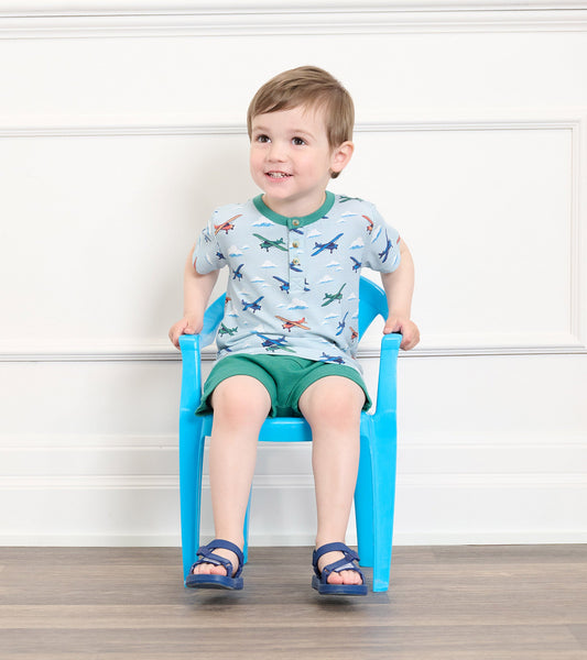 Light blue bamboo henley shirt with airplane print, paired with green shorts, worn by a smiling toddler sitting on a blue ch…