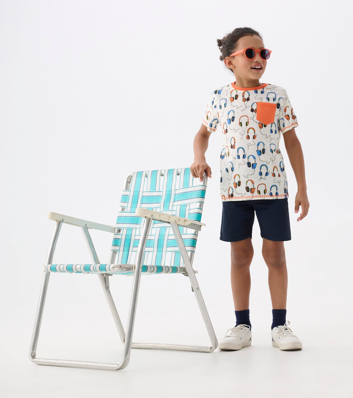 White t-shirt with colorful headphone print and orange pocket, paired with navy shorts, worn by a child.