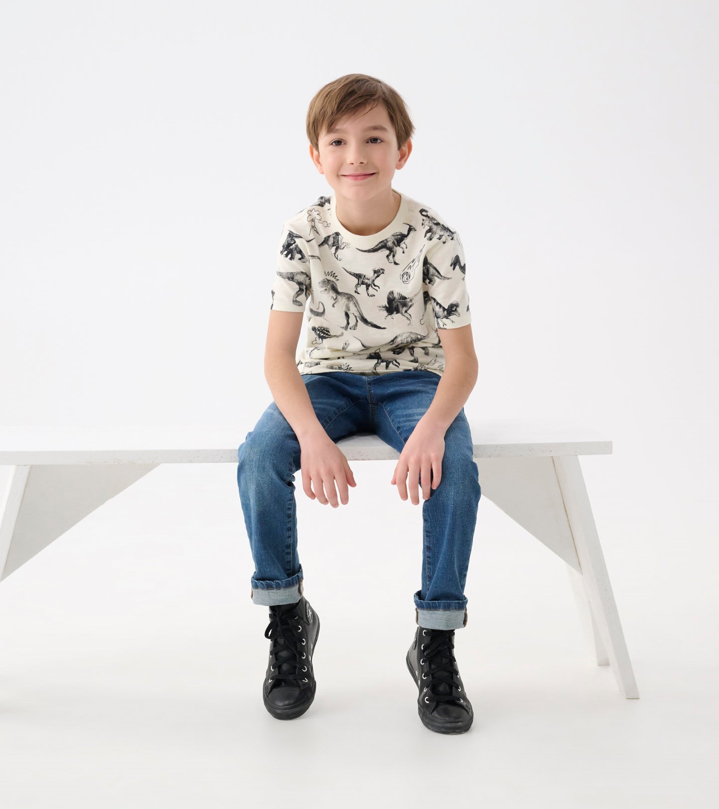 Cream graphic tee featuring black dinosaur prints, paired with blue jeans and black sneakers, worn by a smiling boy.