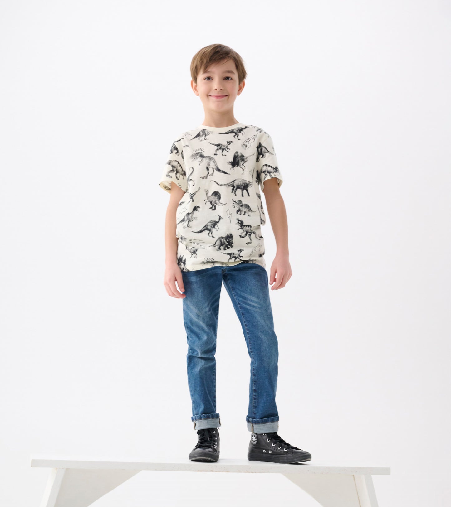 Cream graphic tee featuring black dinosaur prints, paired with blue jeans and black sneakers. Model stands on a white platfo…