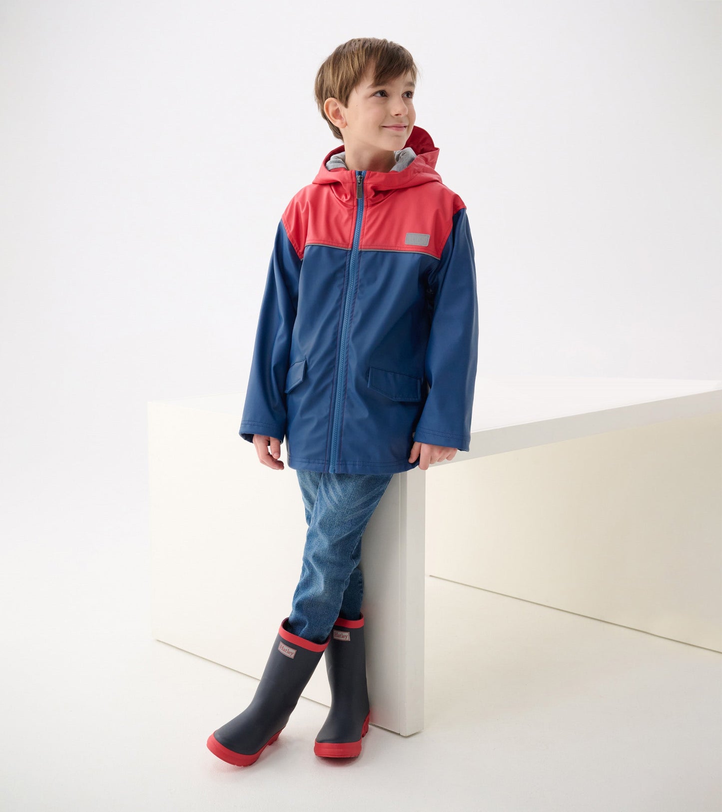 Boys' zip-up rain jacket in red and navy, featuring a hood and two front pockets, worn with jeans and rain boots.