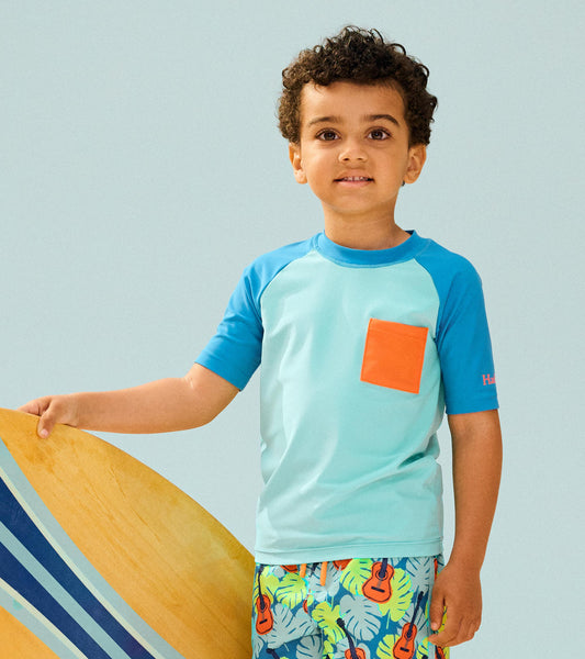 Light blue short sleeve rashguard with orange pocket, worn by a boy holding a surfboard, ideal for beach activities.