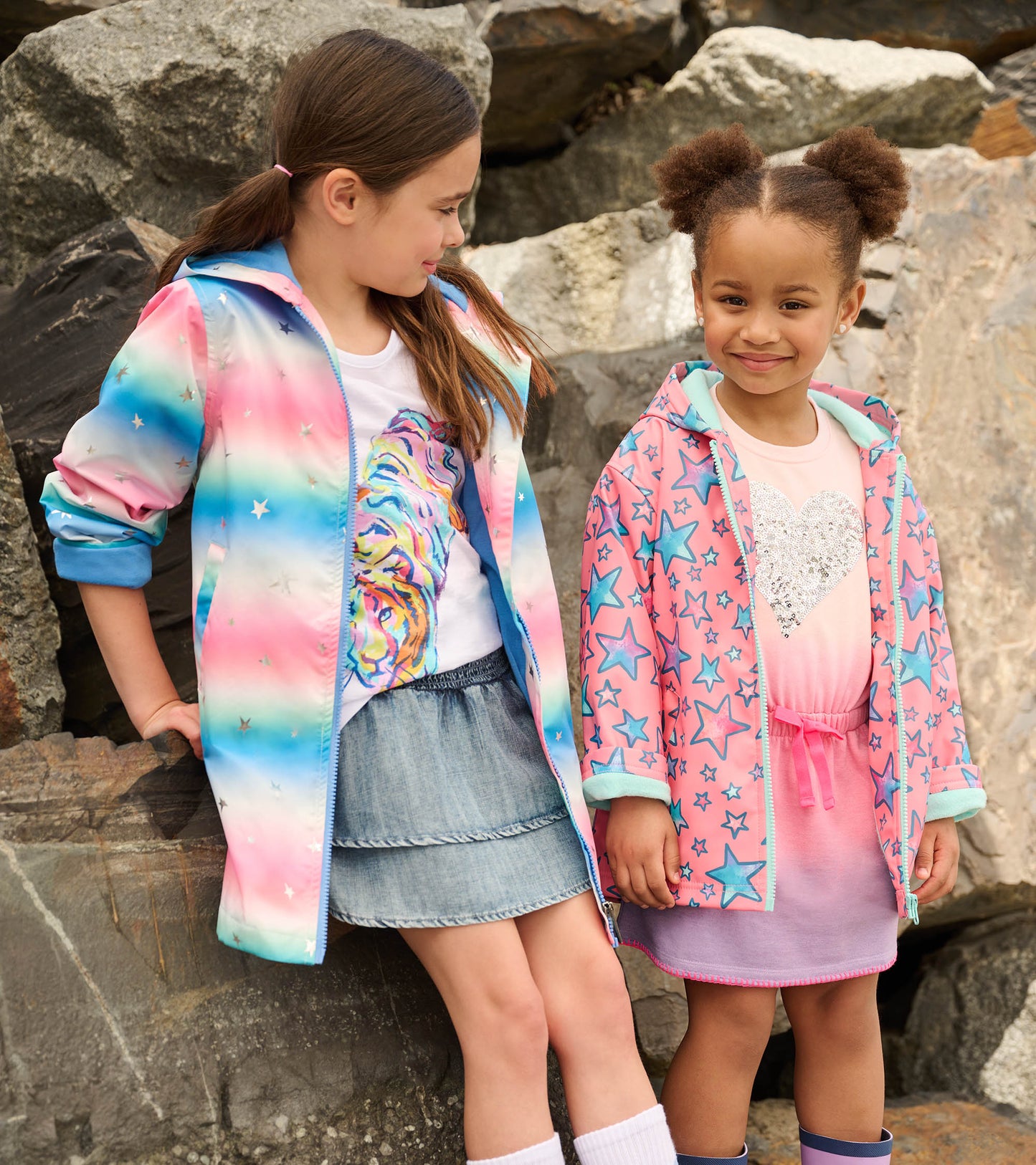 Colorful zip-up rain jackets with watercolor patterns and star designs, worn by two girls against a rocky backdrop.