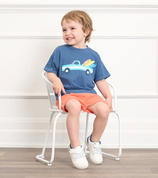 Blue slouchy tee featuring a truck and surfboard graphic, paired with orange shorts, worn by a smiling toddler.