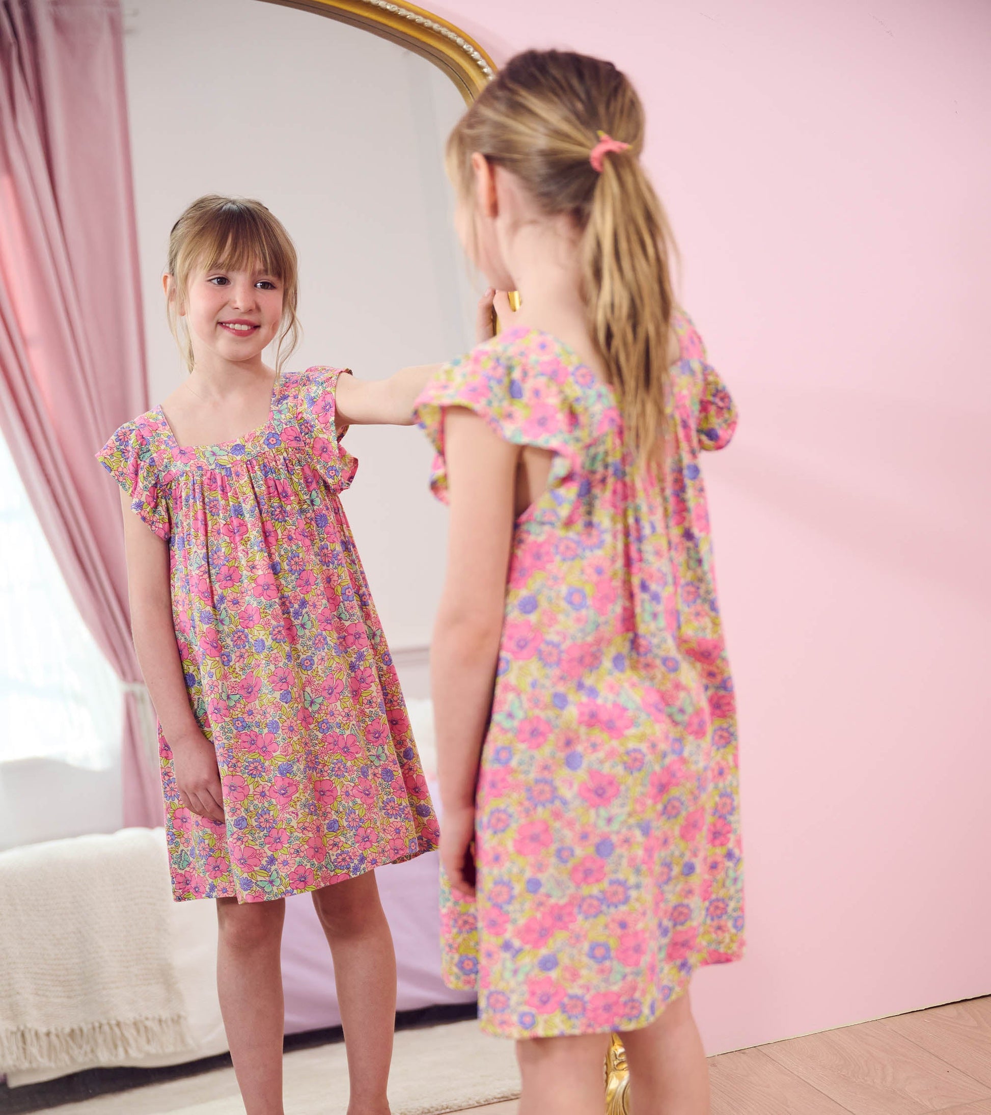 Pink floral dress with flutter sleeves, featuring a relaxed fit, worn by a girl admiring herself in a mirror.