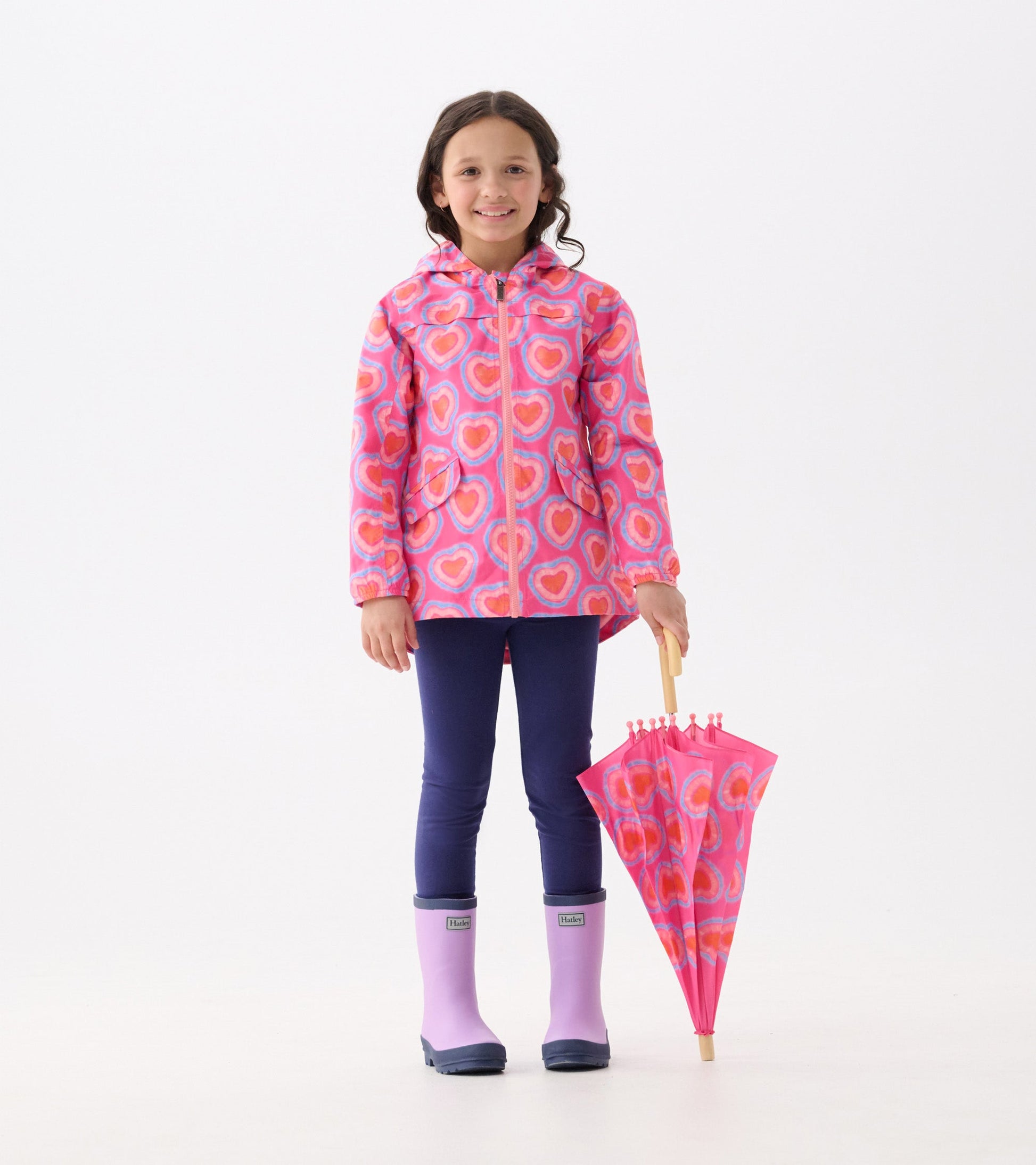 Lightweight pink rain jacket with heart pattern, paired with navy leggings and purple rain boots, held umbrella.