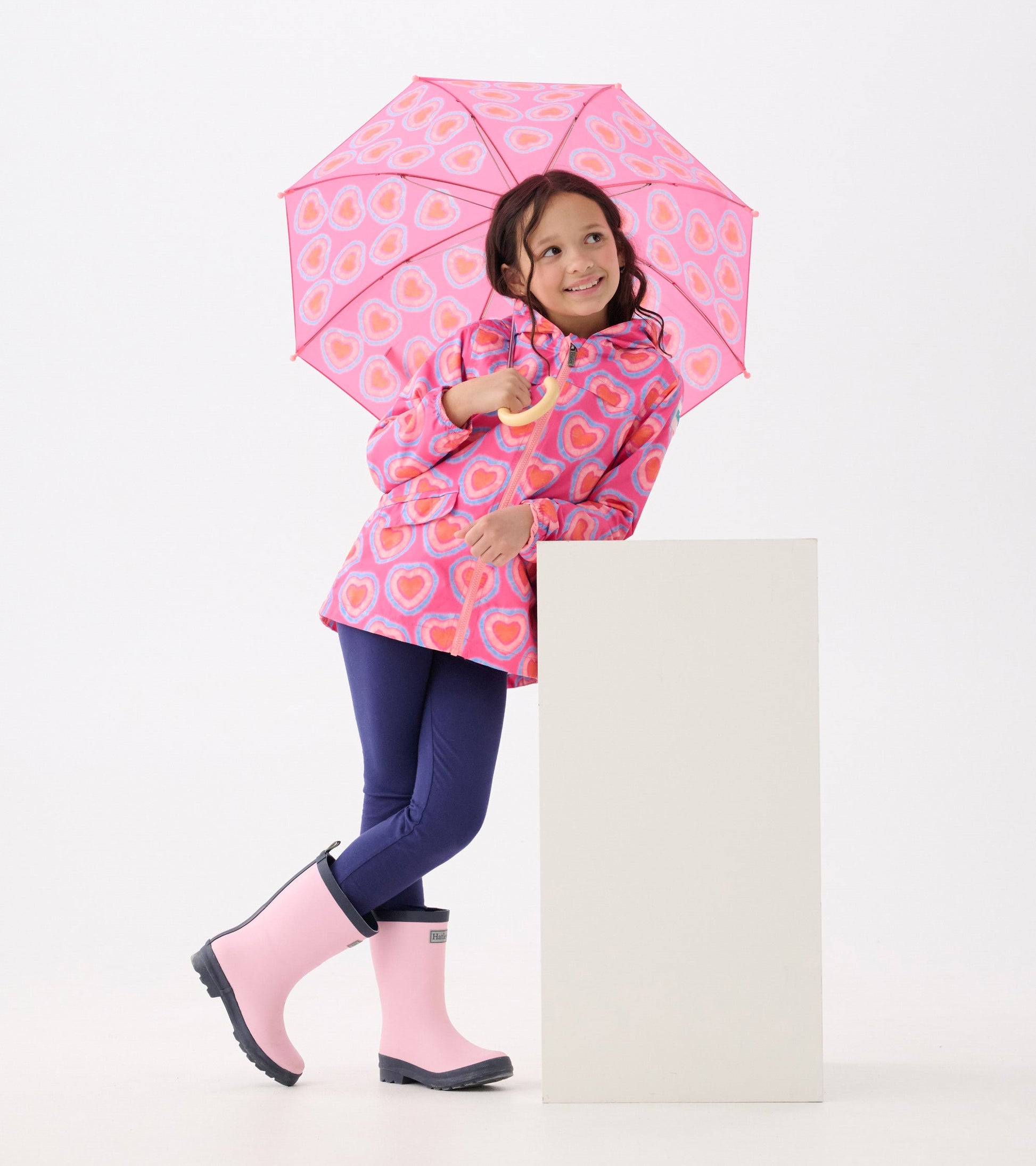 Lightweight pink rain jacket with heart pattern, paired with navy leggings and pink rain boots, held with a matching umbrell…