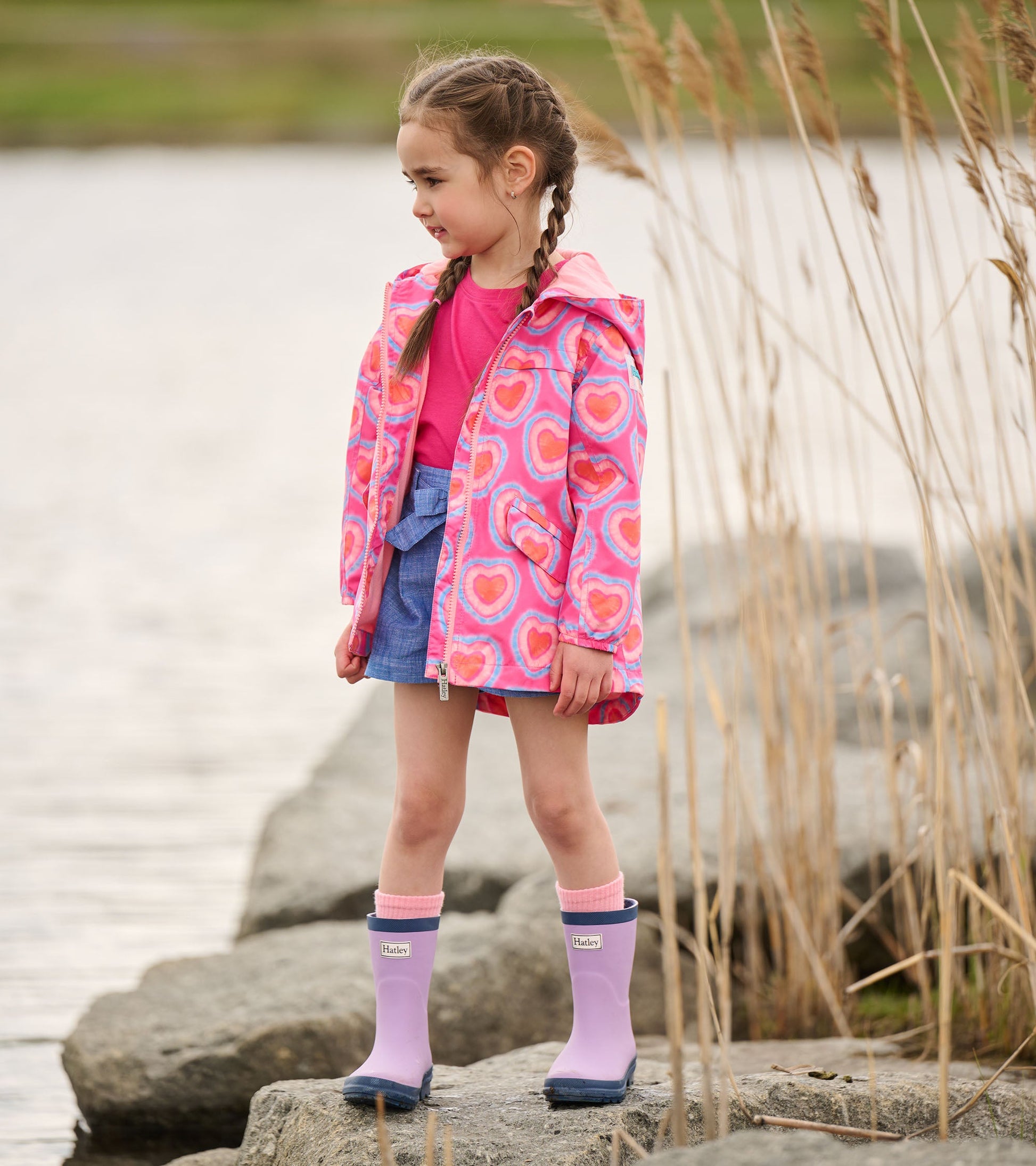 Lightweight zip-up rain jacket with a pink heart pattern, paired with a pink top and denim shorts, worn by a girl outdoors.