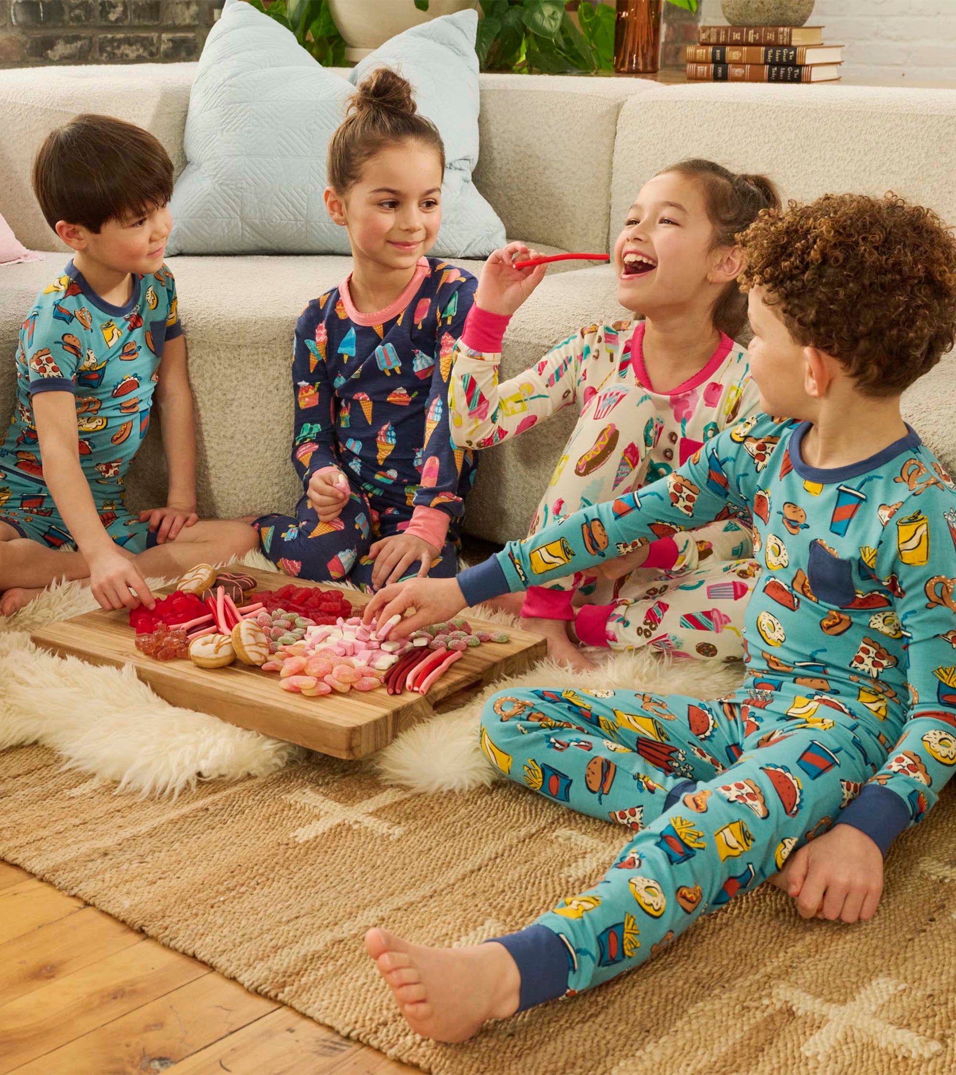 Colorful pajama set featuring ice cream and treat patterns, worn by children sitting on a rug with snacks on a wooden tray.