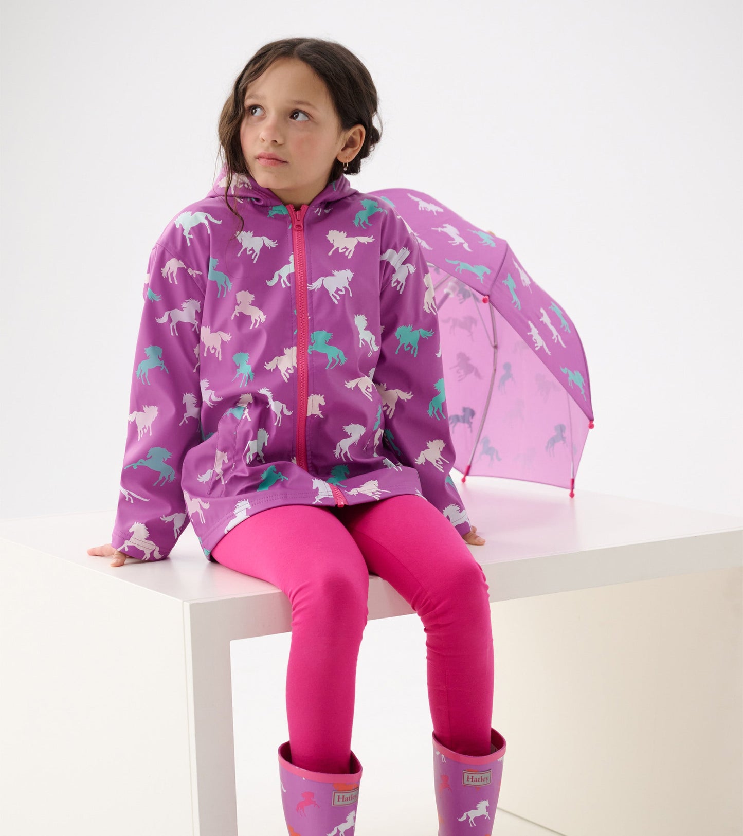 Purple zip-up rain jacket with horse patterns, paired with pink leggings and boots, worn by a girl sitting on a white surfac…