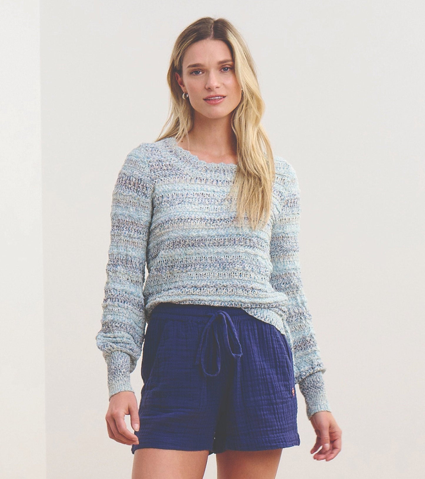 Light blue pointelle sweater with wavy stripes, featuring long sleeves and a relaxed fit, paired with navy shorts.