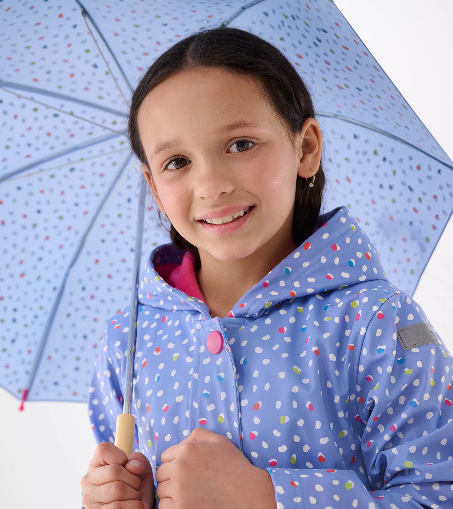 Light blue button-up rain jacket with colorful dot pattern, featuring a hood and pink lining, held by a smiling girl.