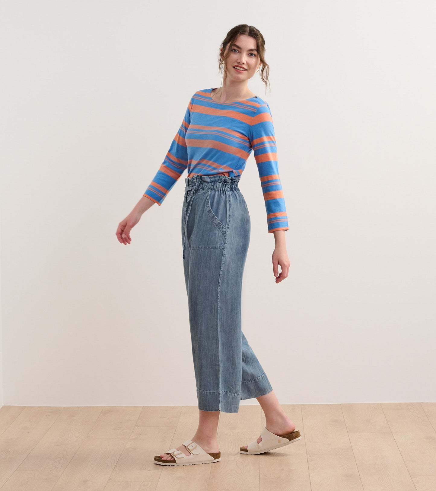 Long-sleeve striped top in blue and melon, paired with light denim pants and sandals, model posing sideways.