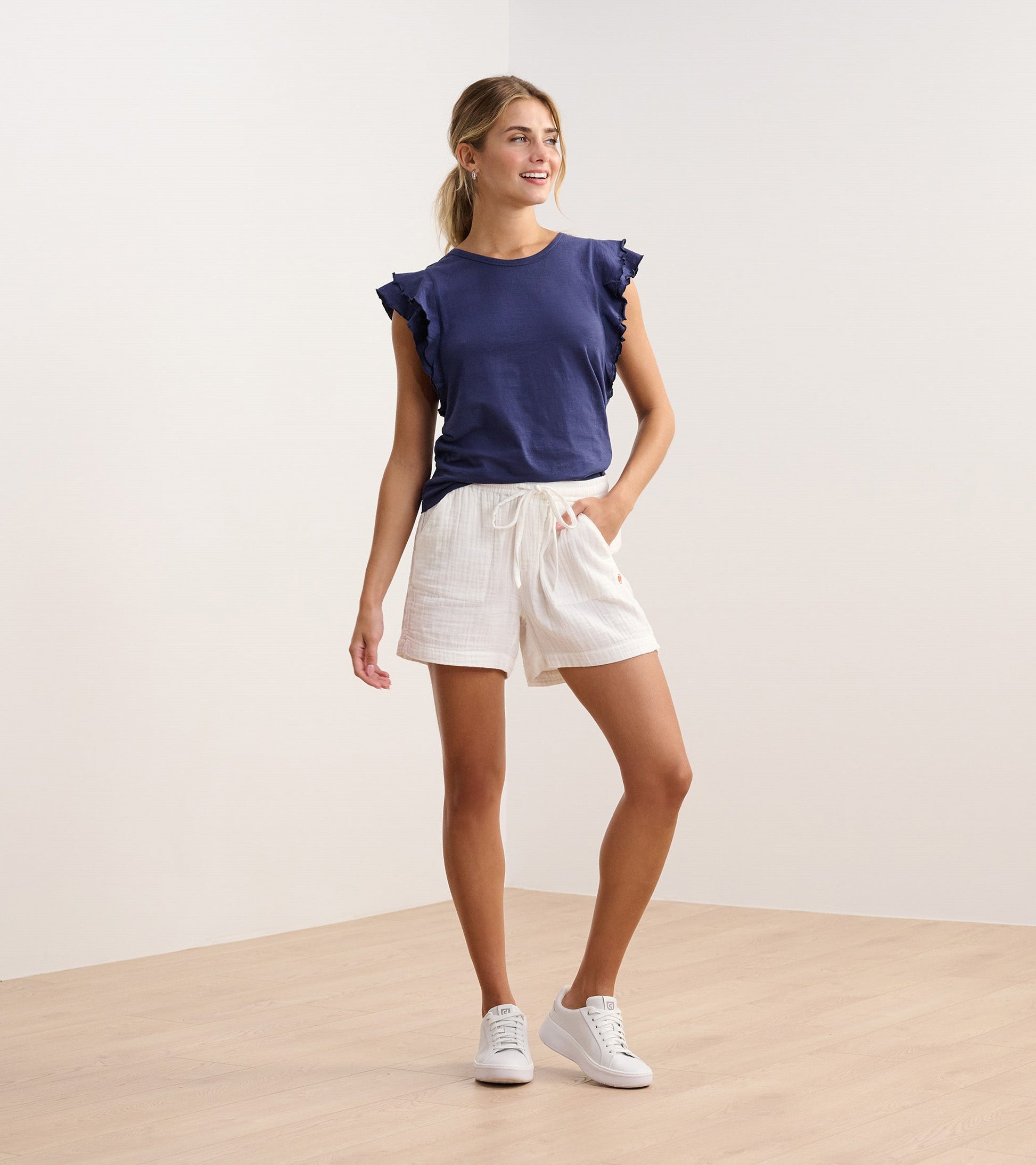 Patriot blue tank top with layered ruffle sleeves, paired with white shorts and white sneakers, standing on a wooden floor.