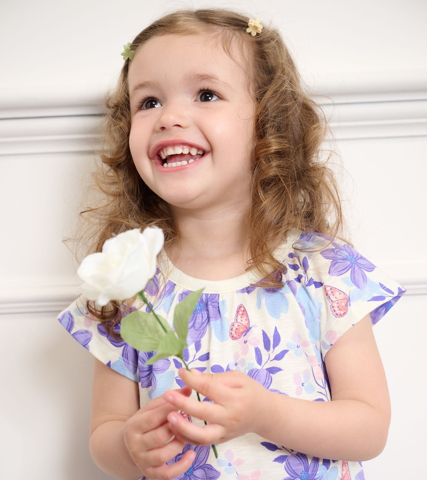 Light yellow dress with purple floral pattern, short sleeves, worn by a smiling girl holding a white flower.