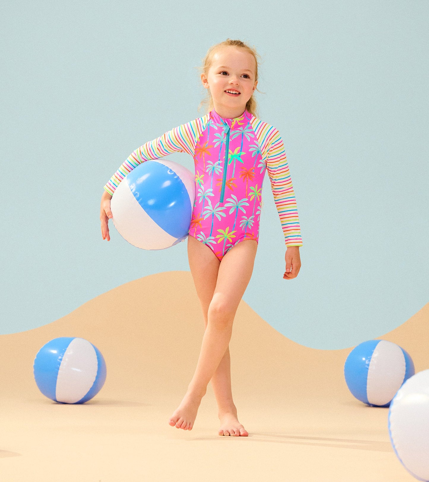 Bright pink long sleeve rashguard swimsuit with palm print, paired with a beach ball, set against a sandy background.