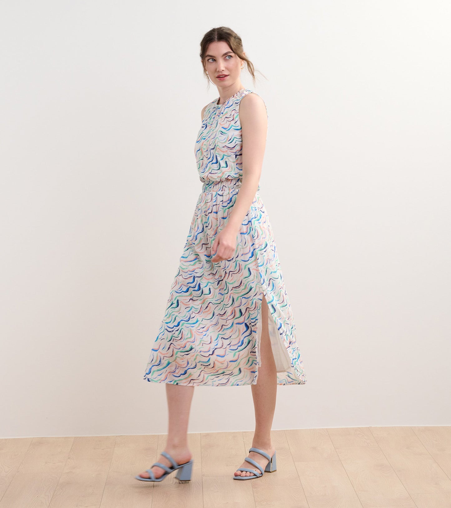 Sleeveless column dress with wavy stripes in pastel colors and a side slit, styled with blue heeled sandals.