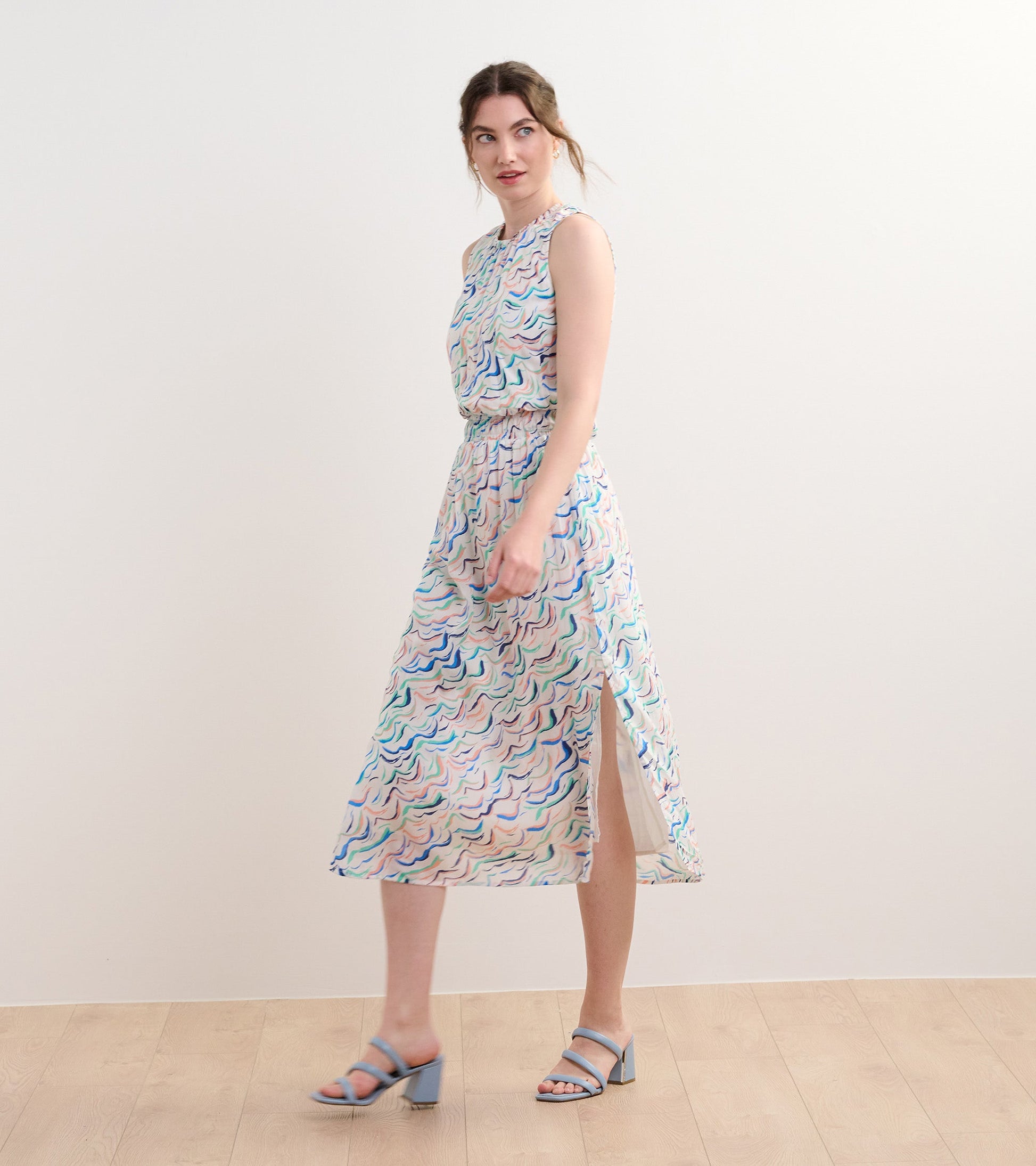 Sleeveless column dress with wavy stripes in pastel colors and a side slit, styled with blue heeled sandals.