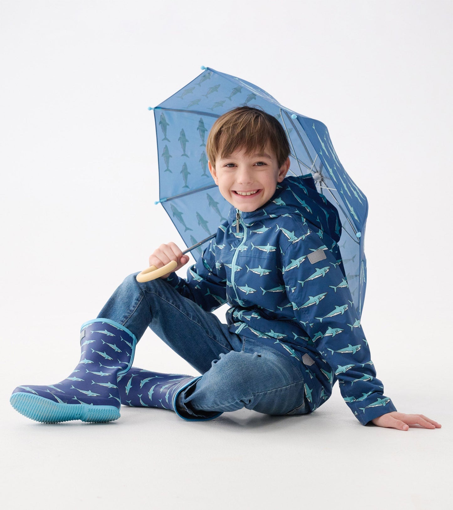 Lightweight blue rain jacket with shark pattern, worn by a boy sitting with a matching umbrella and rain boots.