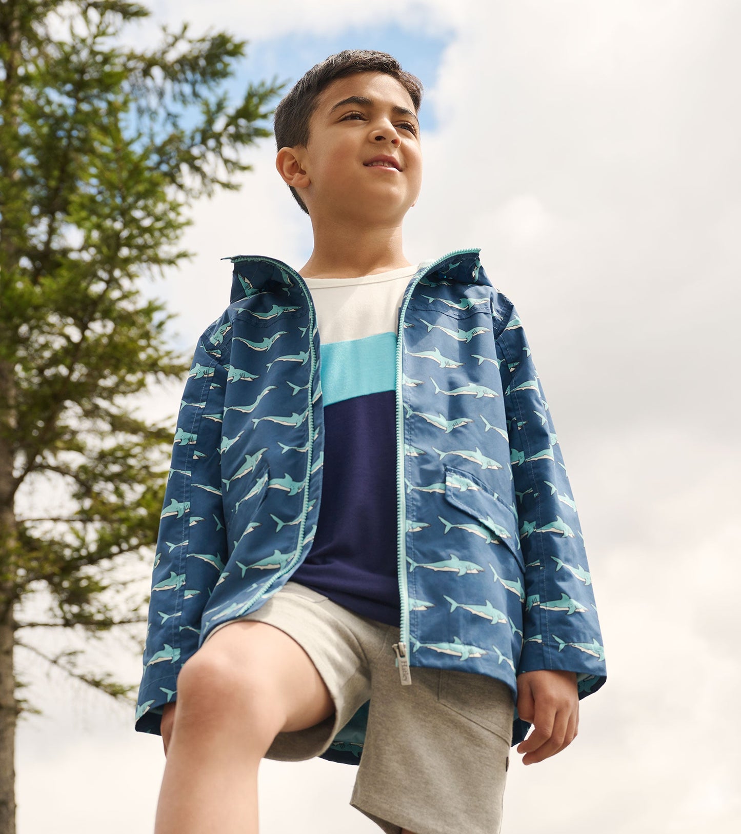 Lightweight blue rain jacket featuring a shark pattern, zip-up front, and hood, worn by a boy outdoors.