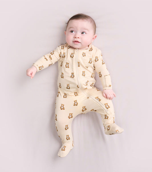 Light beige footed sleeper with teddy bear print, featuring a front zipper and long sleeves, suitable for infants.