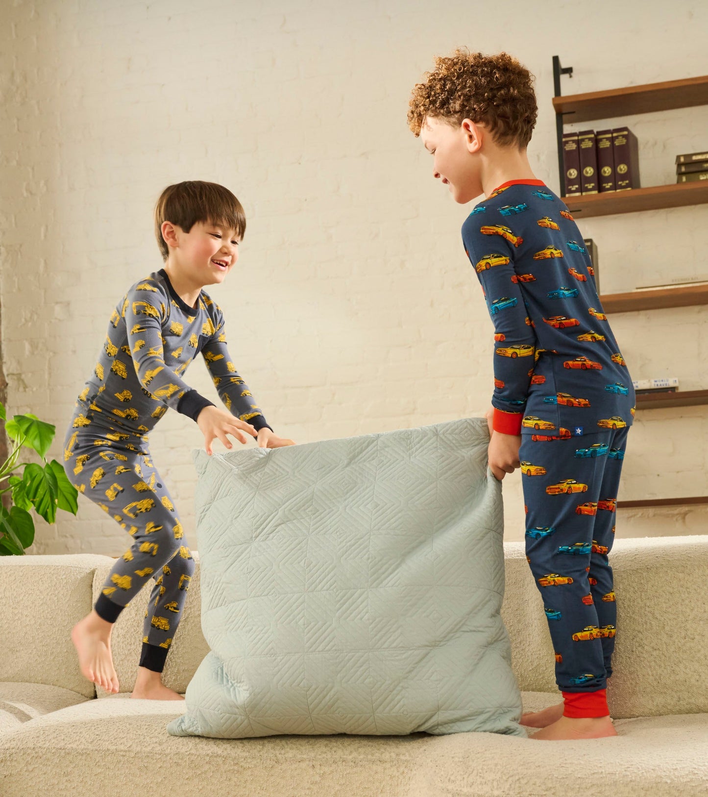 Boys playfully lifting a pillow while wearing colorful car-patterned bamboo pajamas in a cozy living room setting.