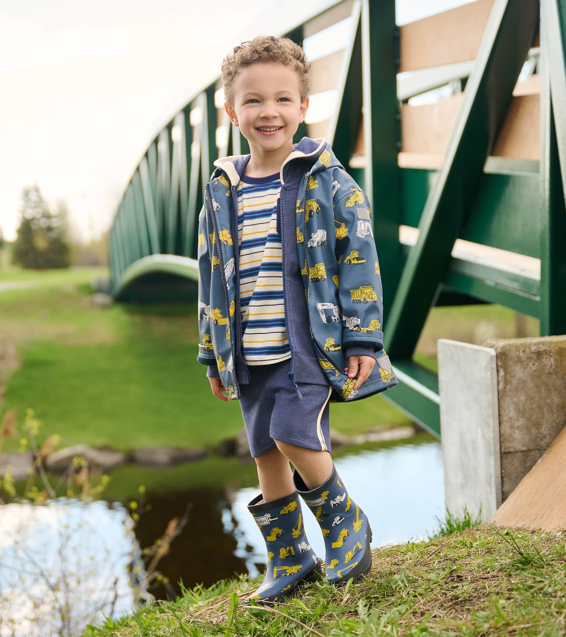 Blue zip-up rain jacket with construction vehicle patterns, worn by a smiling boy outdoors, paired with shorts and rain boot…